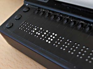 A refreshable braille device sits atop a table. The black, plastic display shows ten braille cells with white four feature brailled characters. 