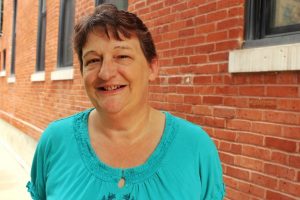 A middle-aged woman with short brown hair and brown eyes wearing a teal shirt with a red brick wall behind her.