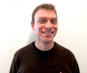  A young man with short brown hair and blue eyes wearing a black sweater with a white background. 