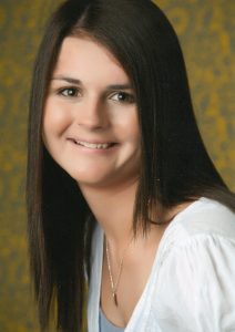  A young girl with brown hair and brown eyes, wearing a white shirt and necklace with a green and yellow splotted backdrop behind her. 