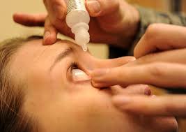 A young brown-haired woman receives eye drops from a staff person at a clinic.