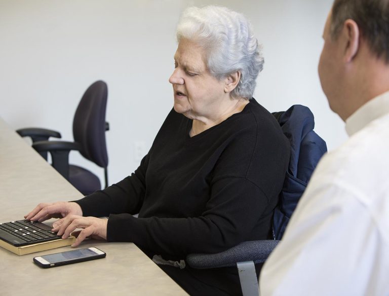 A woman has her hands on a keyboard with bump dots that is hooked up to her iPhone. A man sits next to her