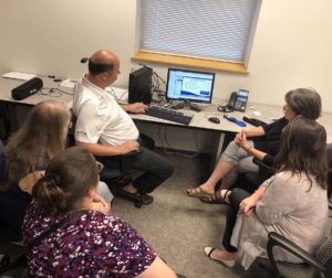 A man sits at a computer screen and a group of people sit around him.