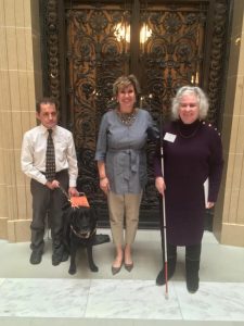 Three people stand in Capitol building. One has a white cane and one has a service dog.