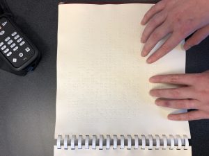 Two hands reading brailled text of a recipe in a cookbook. A black Trek reader is also on the table.
