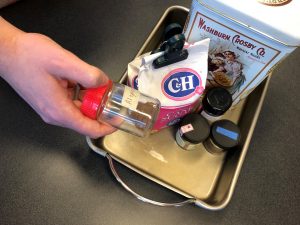 A hand places an small container marked “allspice” into a baking pan. The grey metal baking pan contains a tin of flour, bag of sugar and other small spice containers.