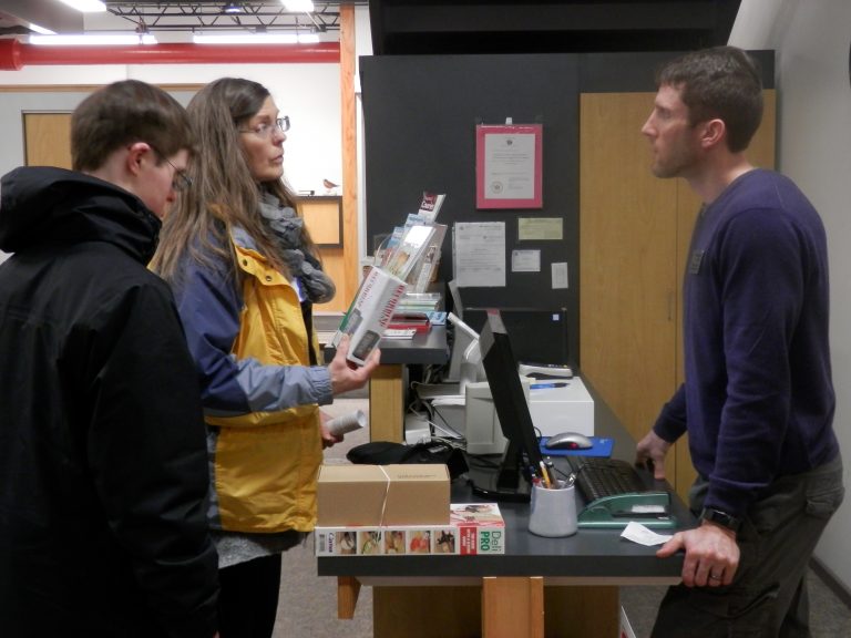A man behind a cash register talks to a woman and her son. The mother is holding a store product.