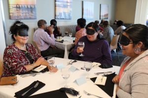 Three women who are blindfolded sit at a table.