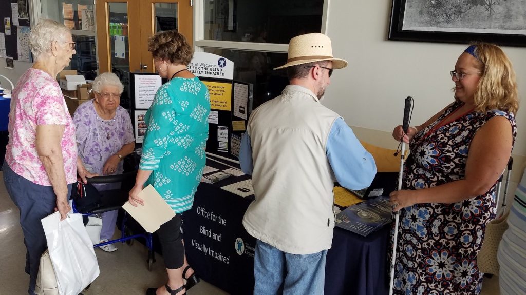Five people stand in front of a vendor table. One has a walker and one has a white cane.