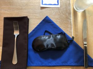A place setting with a blue napkin folded in a triangular shape and a blindfold resting on top of it.
