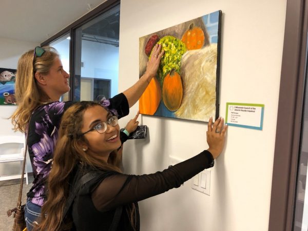 A woman feels a painting while a girl with thick glasses holds the painting steady on the wall.