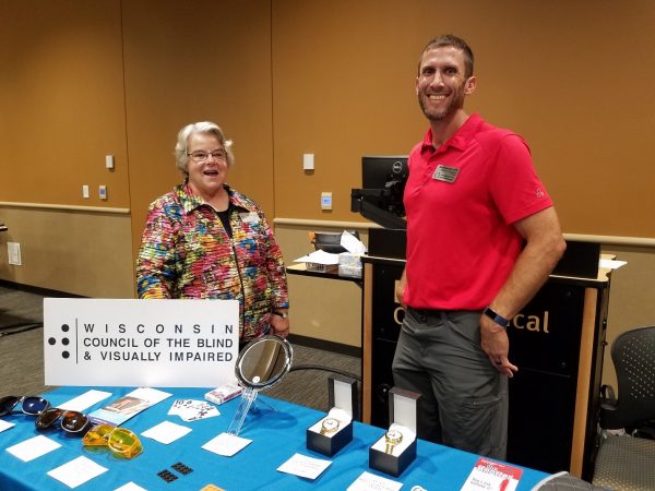 Two people stand at a vendor table with the Wisconsin Council of the Blind & Visually Impaired.