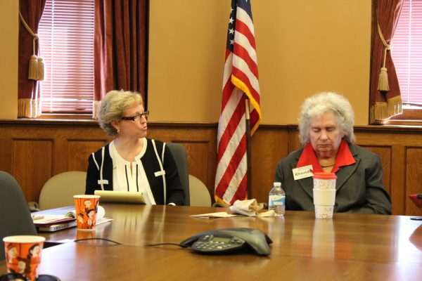 Two women sit at a conference table with an American flag behind them.