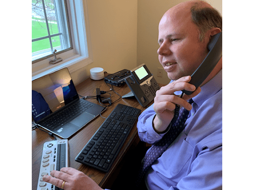A man on the phone with a braille keypad and keyboard in front of him. 