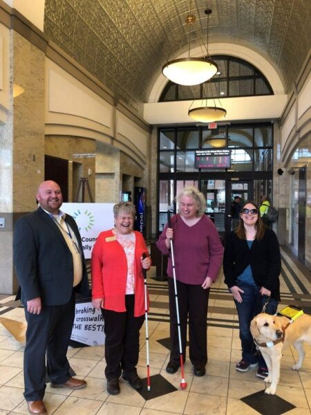 Four people stand in a public transit building; two have white canes and one has a service dog.