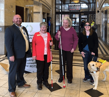 Four people stand in a public transit building; two have white canes and one has a service dog.