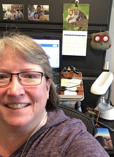 A woman smiles at the camera with a laptop on a desk in the background.