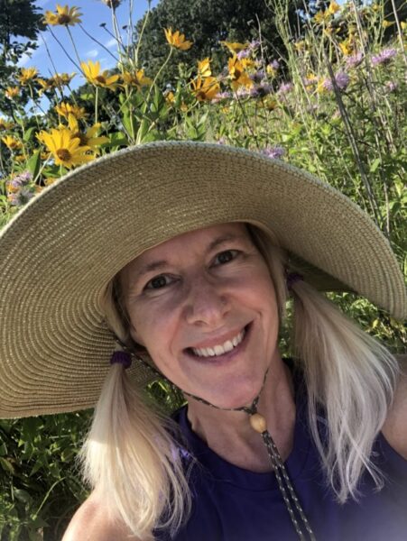A woman with a large gardening hat smiles in a selfie with a garden behind her.