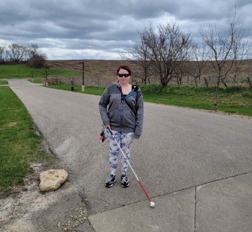 A woman stands holding her white cane on pavement.