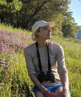 A woman with binoculars sits in a meadow as she listens to birds.