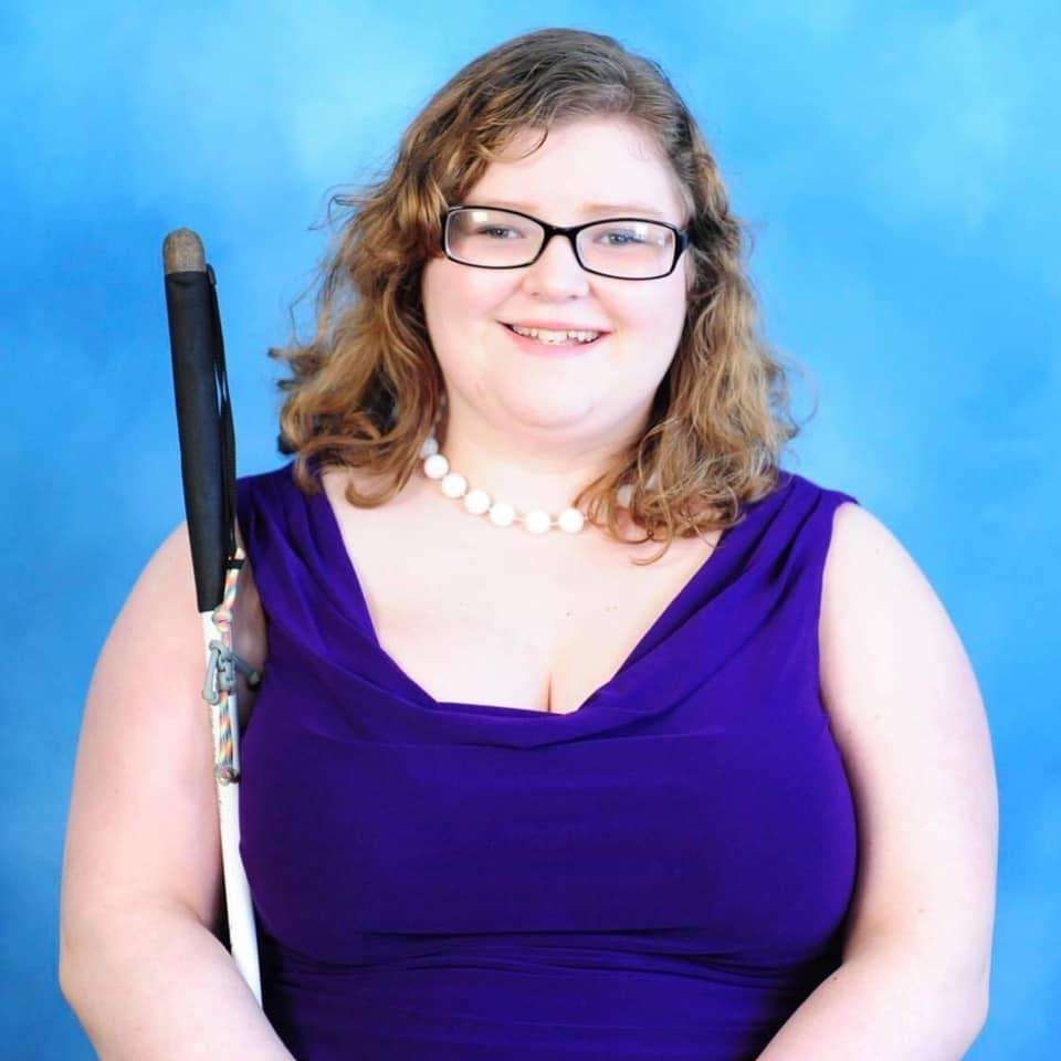 A young woman with glasses wearing a purple dress holding a white cane.