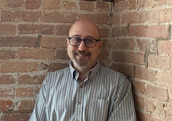 A bald man wearing glasses standing in a corner formed by brick walls.