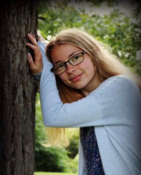 A young woman with glasses wearing a sweater leaning her head on her hands against a tree.