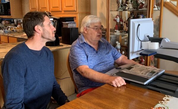Photo of two men sitting side-by-side at a table, the one on the right using a video magnifier to read a newspaper.