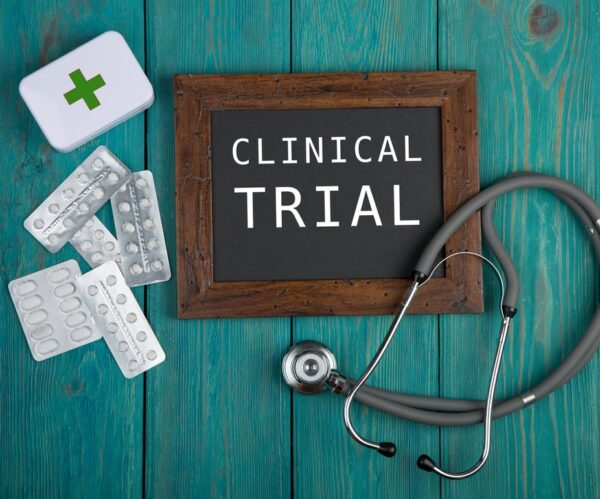 Photo of a framed sign reading Clinical Trial, a stethoscope, some pills and a white box with a green cross on it.