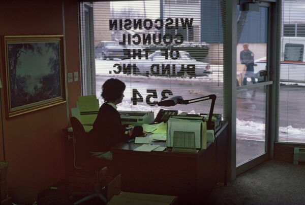 A woman sits at a desk with a large window behind her.