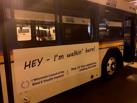 A parked Wausau Metro Ride bus displaying a large ad on the side reading “Hey—I’m walkin’ here! Stop 10’ from pedestrians crossing the street” along with the Wisconsin Council of the Blind & Visually Impaired logo.