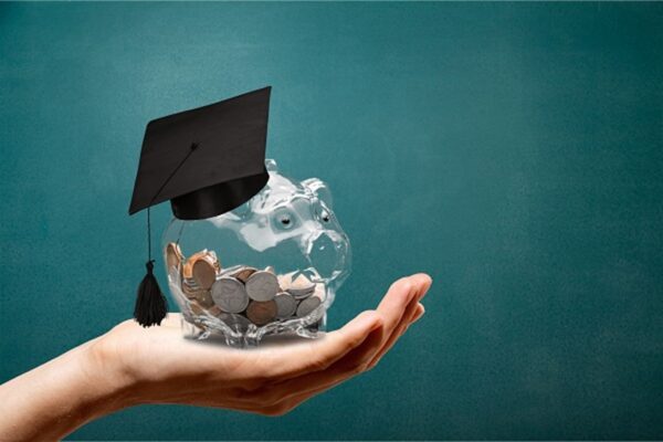 A hand holding a clear piggy bank with a mortarboard on its head