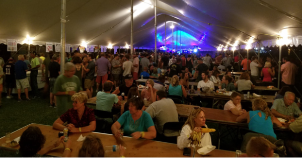 A crowd of people in a large festival tent eating corn and brats