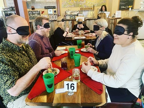 Blindfolded diners sitting at a long table eating a meal