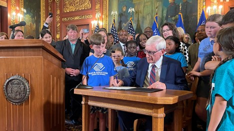 Governor Tony Evers sitting at a desk signing the 2023-25 State Budget as a crowd of people stands behind him watching