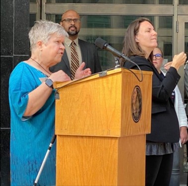 Denise Jess standing at a podium speaking at a press event