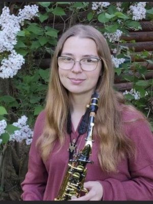 Grace Cain holding a saxophone and standing in front of flowering foliage