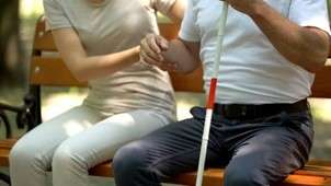 Two people sitting on a park bench, one of them holding a white cane and the other offering assistance
