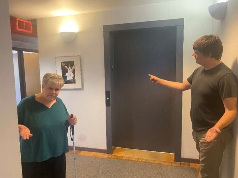 A sighted person attempting to direct a person with a white cane toward an elevator by pointing at it.