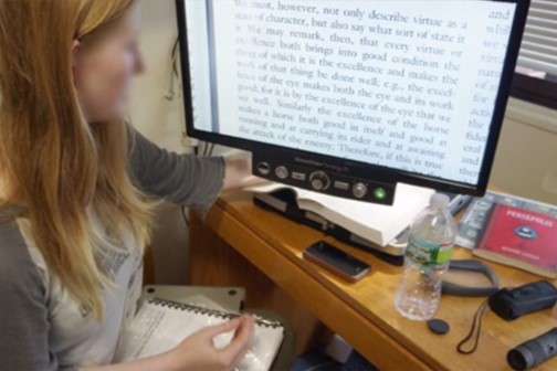 A woman sitting at a desk using a video magnifier to read a textbook.