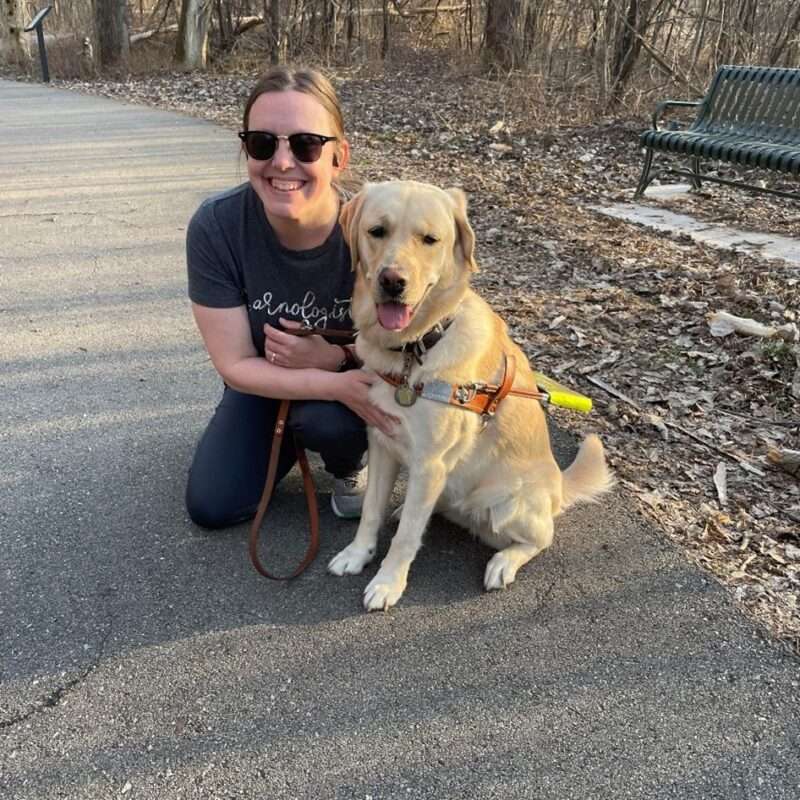 Liz Fryseth kneeling on a paved path with her guide dog.