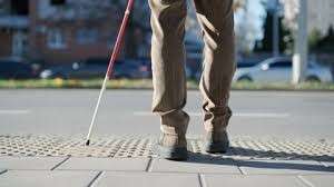 The feet of a person with a white cane standing at a curb.