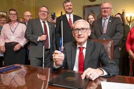 Gov. Tony Evers sitting at a table signing a bill with a group of advocates standing behind him