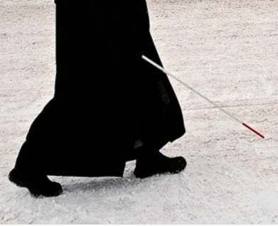 The legs of a person walking on snow with the aid of a white cane.
