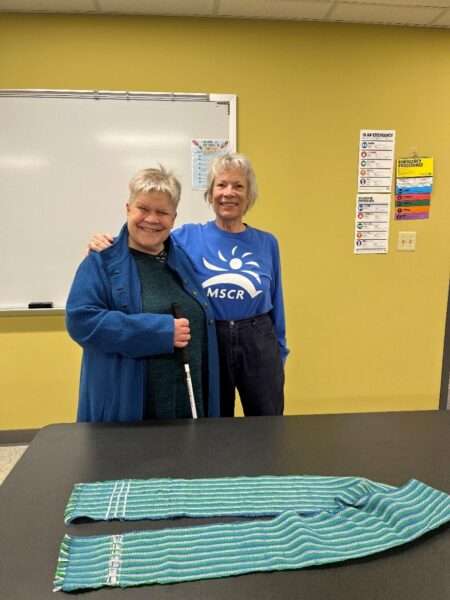 Two women standing behind a table on which a woven scarf is displayed.