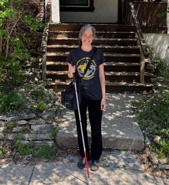 A light-skinned women holding a white cane standing in front of porch steps