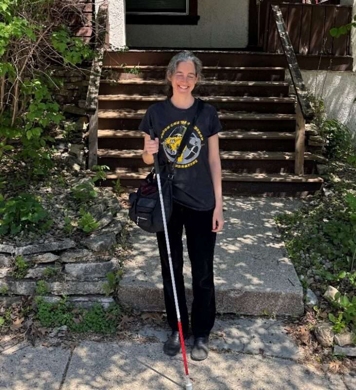 A light-skinned women holding a white cane standing in front of porch steps
