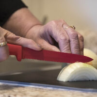 Close-up of a woman’s hands chopping a yellow onion using a red knife.