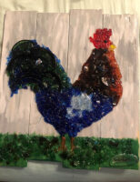 A rooster made of crushed glass stands on a bright green glass grass; four white boards make up the background of this piece.